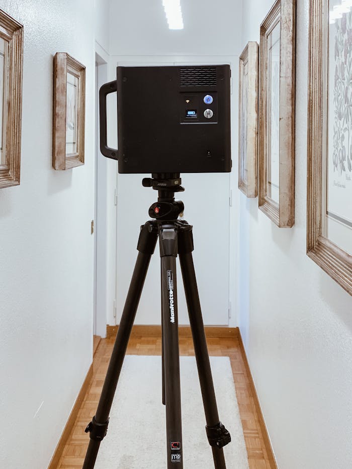 A tripod-mounted scanner in a bright corridor enhances the artistic appeal of framed paintings on the walls.